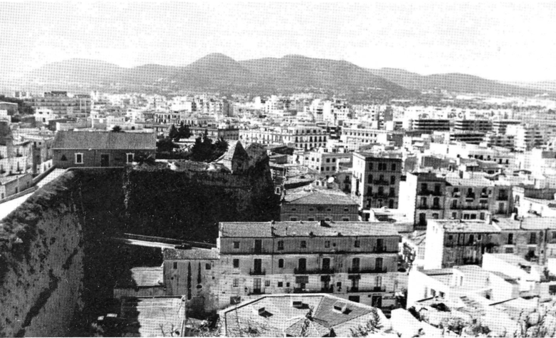 A vol d'ocell des del baluard de Santa Llúcia tenim una nova perspectiva de la ciutat. En primer terme, la plaça del Carbó amb el terrat poligonal del Mercat de la peixateria i carnisseria vella, coetani del Mercat Vell, el decimonònic «Poble Nou» de la Marina i, al centre de la fotografia, l'eixampla de Vara de Rey. Més enllà, en un desordre aparent, la recent expansió urbana, que cobreix gairebé tot el terme municipal, sembla formar un nou mur de ciment delimitat pel cinturó de ronda. Al fons, els puigs de la sèrie tectònica d'Eivissa.