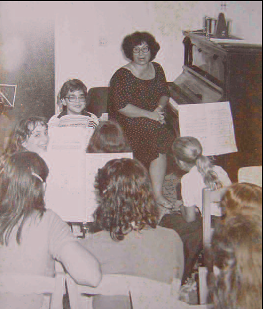Una classe de solfeig. La fotografia correspon al mes d'octubre de l'any 1980, quan l'escola ja havia passat a convertir-se en aula d'extensió del conservatori. Tot i això, il·lustra perfectament la manera de treballar de quan encara era escola: els alumnes asseguts en cadirons encordats, sense ni tan sols taula, al voltant de Lina Bufí, qui els acompanyava amb el piano per cantar les lliçons (fotografia de l'arxiu particular de Lina Bufí).