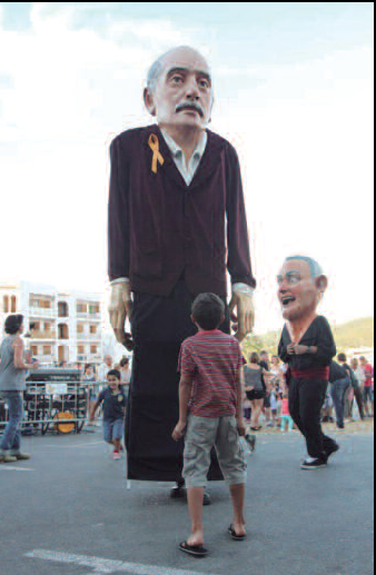 Figura d'un home gran amb una banda groga i vermella al coll, observant una festa popular amb altres figures i gent al fons.