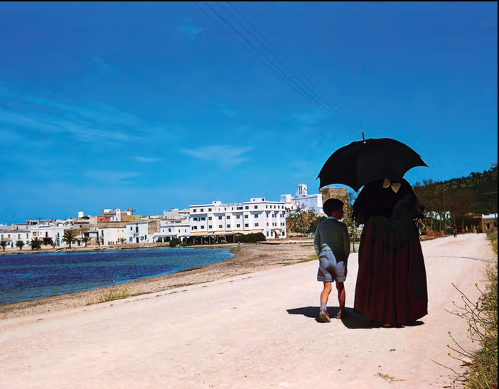 Una pagesa passejant amb un nen per la platja de s'Arenal a Sant Antoni quan el turisme ja estava plenament implantat -es veu l'hotel San Antonio ja acabat-, però no s'havia iniciat el boom de la construcció. Autor Antonio Campañá, 1960. Procedent del llibre de Martin Davies (ed.) Eivissa-Ibiza: L'illa d'un temps (Barbary Press, 2005), pàg. 226.