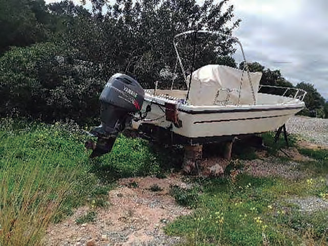 Una petita barca de motor blanca amb un motor fora borda Yamaha està a terra sobre un remolc, en una zona de gespa seca al costat d'un mur de pedra.