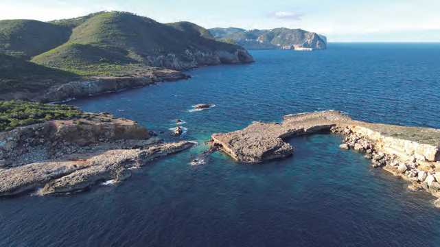 Perspectiva de la costa de la venda de Benirràs amb l'illa d'Encalders en primer terme (dreta) separada de la plana de s'llla (esquerra) pel pas de s'llla. Al darrere, la punta des Catiu i seguidament el vessant costaner del puig des Guixar amb els tallserrats de Davall es Puig, la cova de s'Orenga i la punta del mateix nom (fotografia d'Iñaki de Eróstegui Aznar).